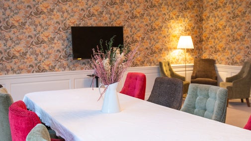A white table with red, green and brown chairs in a room with orange and green floral wallpaper. A TV screen is mounted on the wall and three green chairs are in a cluster in the corner around a table and lamp.